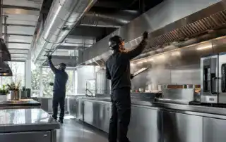 Fotorealistisches Hero-Bild einer modernen Edelstahl-Großküche mit industrieller Lüftungsanlage und sichtbaren Abluftkanälen. Zwei Techniker prüfen Dunstabzugshaube und Filtersysteme in hygienischer Gastronomieumgebung. Fokus auf Küchenabluft, Luftqualität, Wartung und technische Sicherheit nach VDI-2052-Standards.